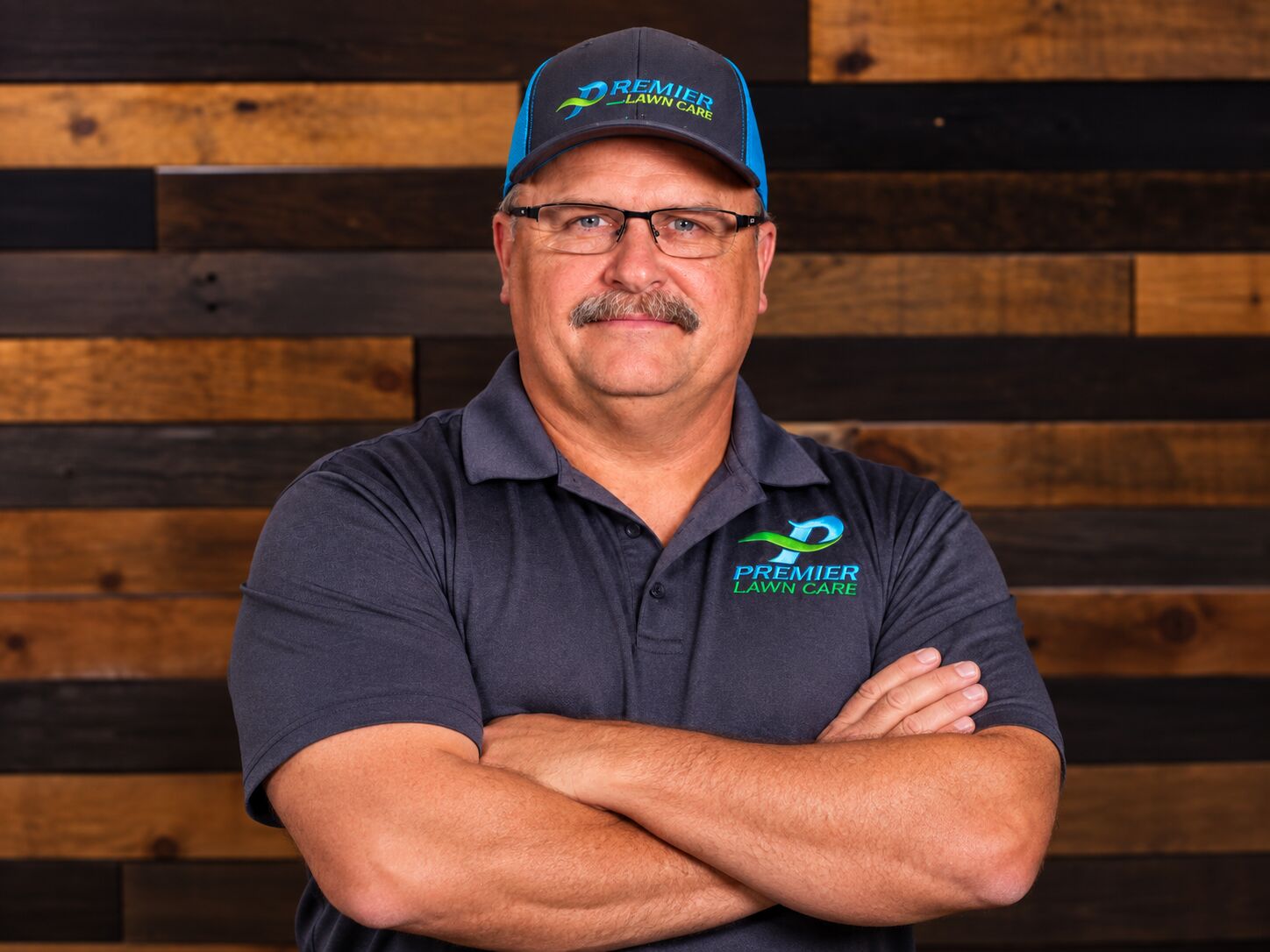 Dennis Herring, President of Premier Lawn Care, standing with arms crossed in a branded cap and shirt