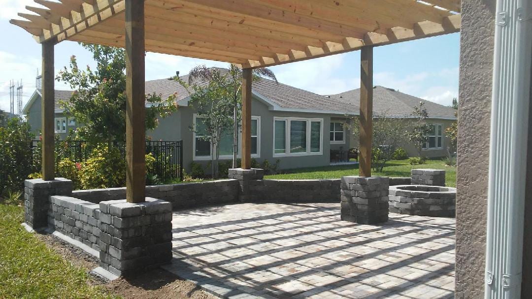 A wood pergola over a paver patio with a stone seat wall and built-in fire pit