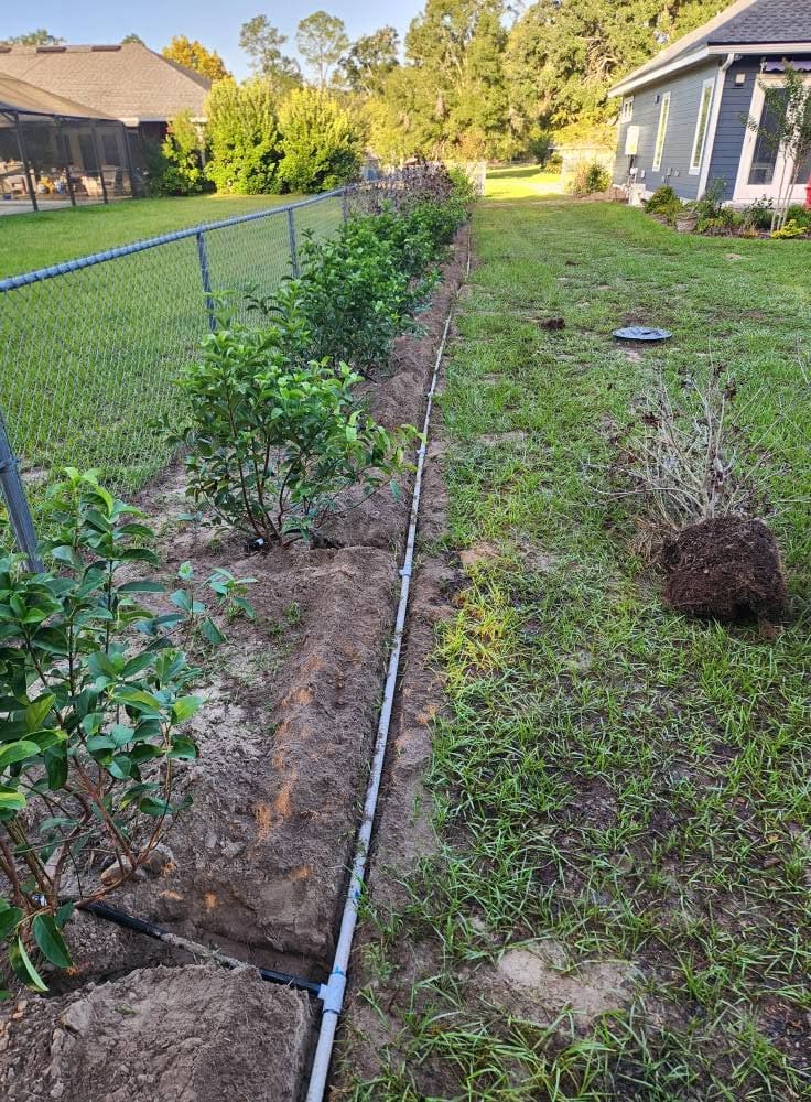 Trenched irrigation pipe running along a fence lined with shrubs in a backyard