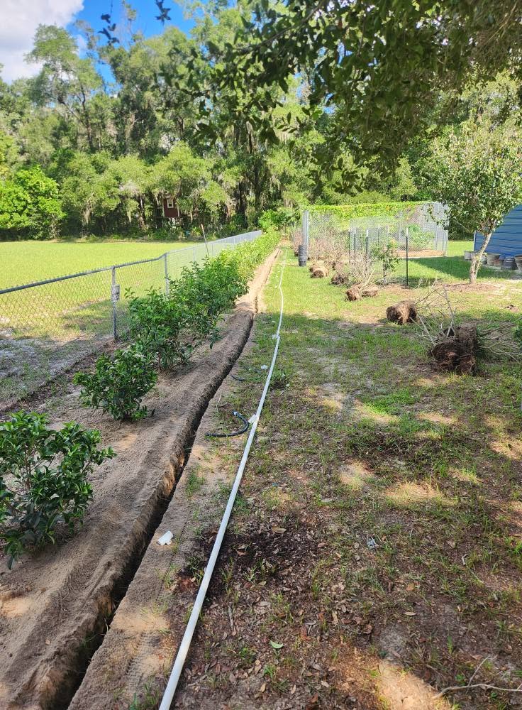Open trench beside patchy grass, with a light-colored pipe and chain-link fence.