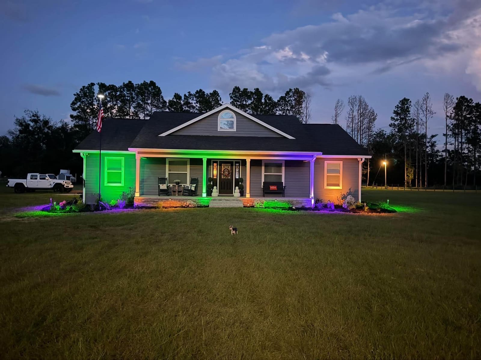 A farmhouse-style home with colorful green, purple, and orange landscape lighting at dusk