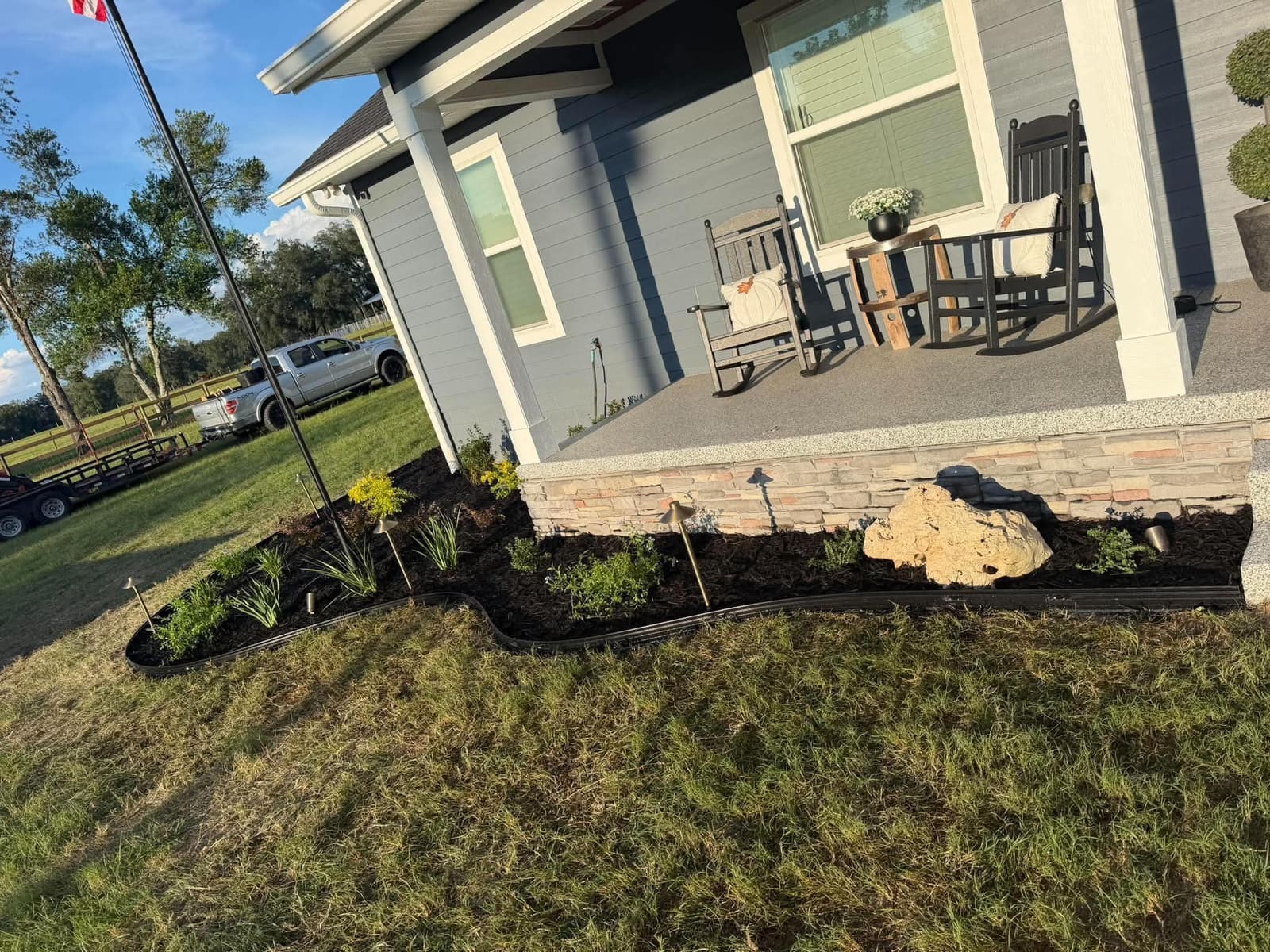A curved mulch planting bed with a large boulder accent and landscape lighting fixtures beneath a covered front porch