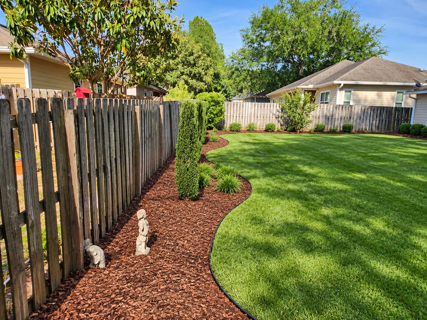 A tidy mulch bed with a kneeling garden statue along a backyard fence