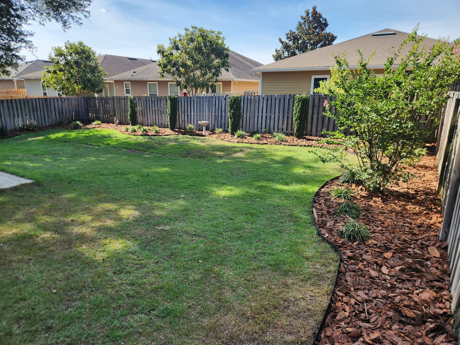 A curved mulch bed with a birdbath and shrubs along a backyard fence