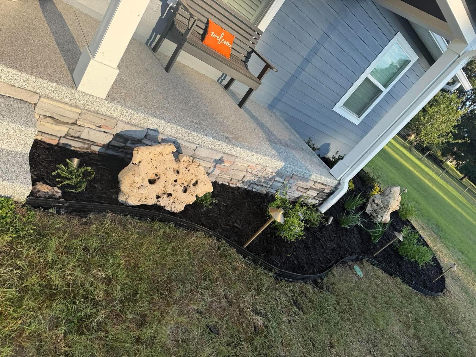 A close-up of a mulch planting bed with a large boulder accent and landscape lighting fixtures near porch steps