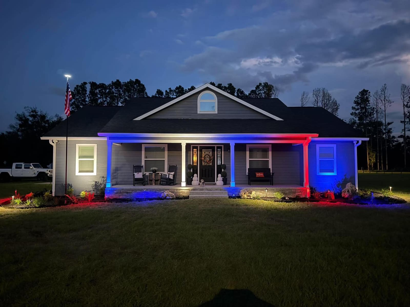 A farmhouse-style home with red, white, and blue patriotic landscape lighting at dusk