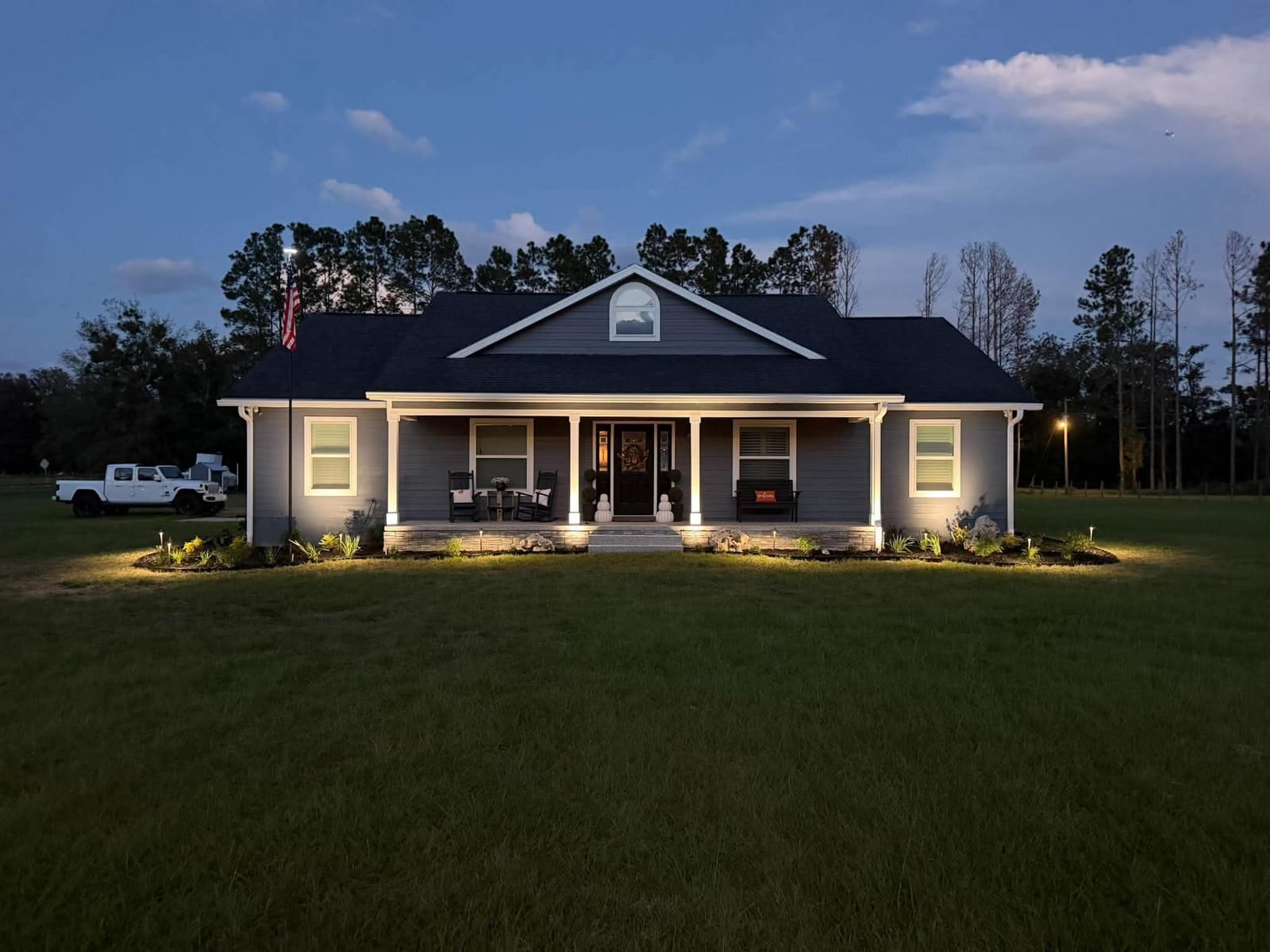A farmhouse-style home with warm white landscape lighting along the porch and roofline at dusk